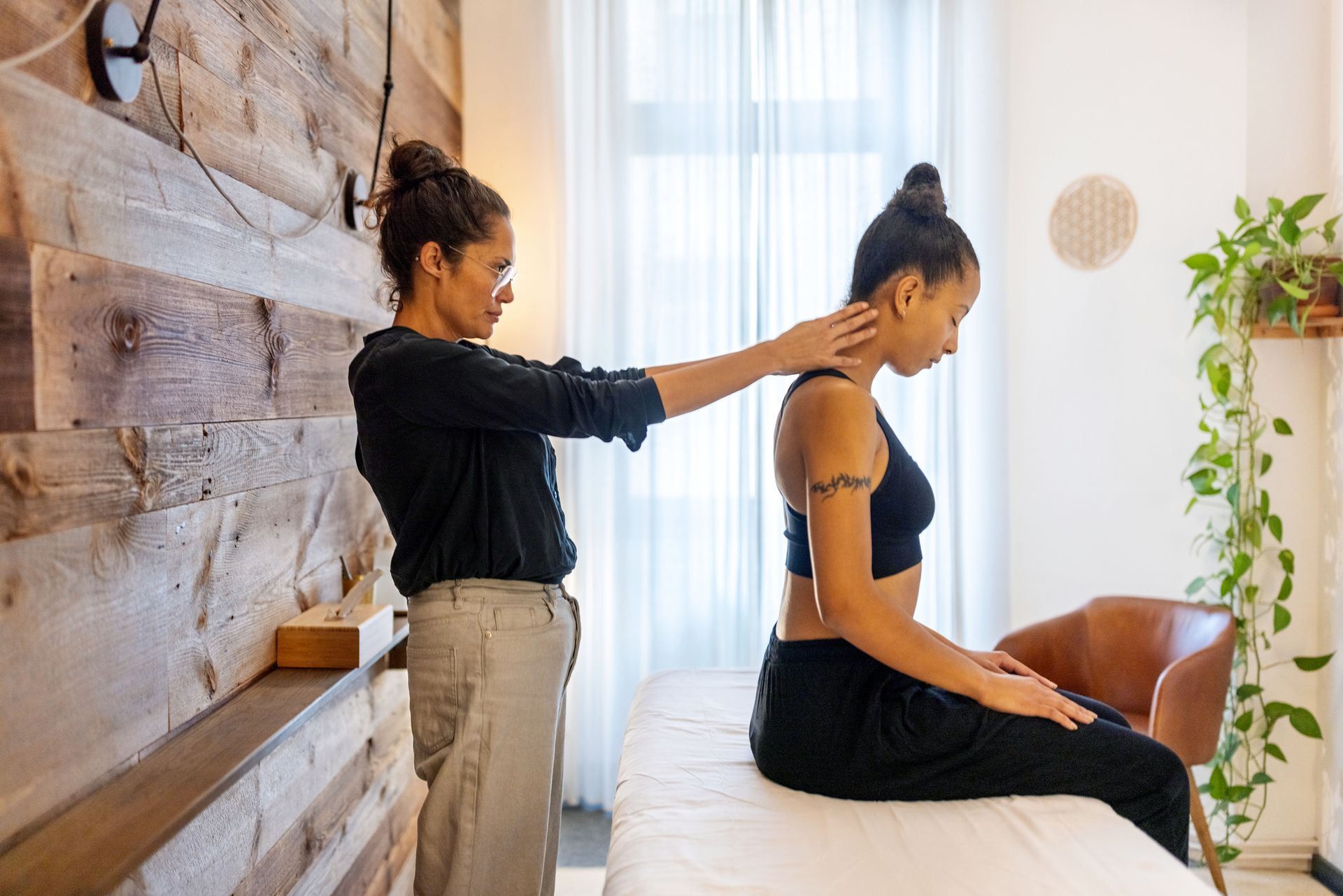 Woman performing a neck massage on another woman seated on a massage table. Wooden wall backdrop.