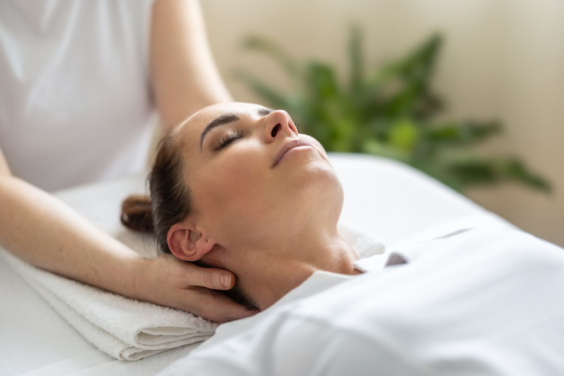 Woman receiving neck massage, lying on a massage table with eyes closed, in a well-lit room.