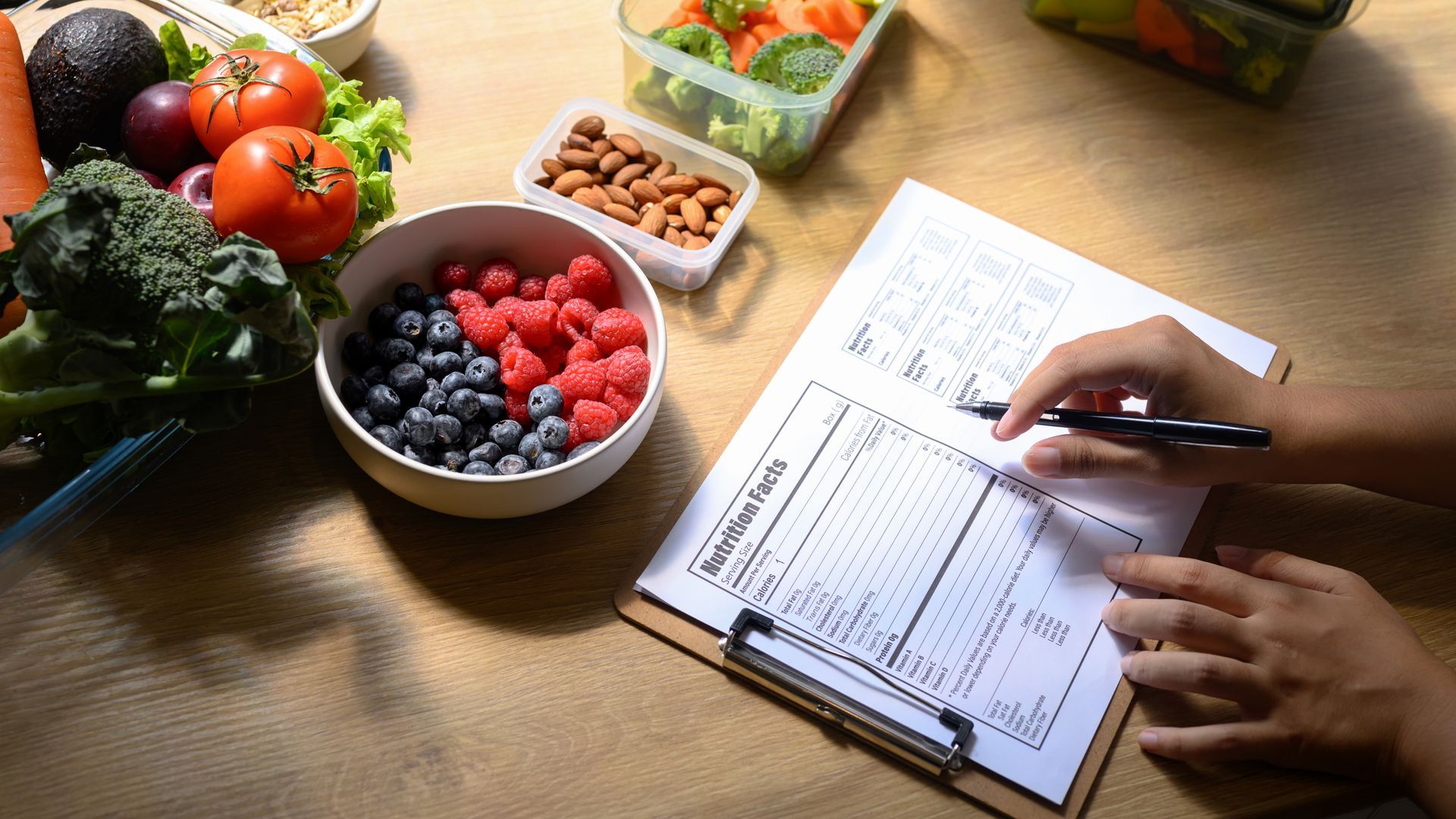 Person writing on a nutrition label with fresh fruits, vegetables, and nuts. Person writing on a nutrition label with fresh fruits, vegetables, and nuts.