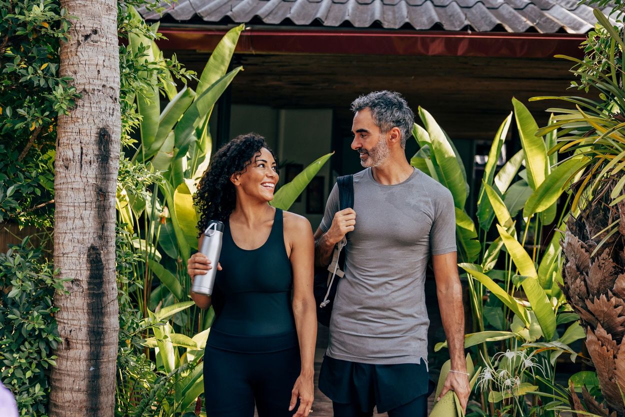 Couple walking together, smiling, near foliage. Woman holds water bottle, man carries backpack and towel.