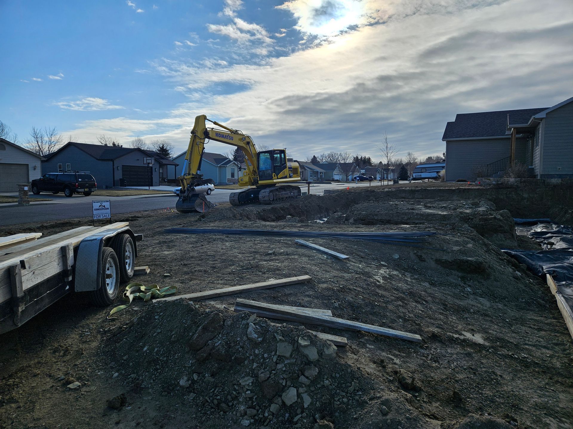 Yellow excavator on a dirt lot. A trailer is in the foreground, houses in the background under a cloudy sky.