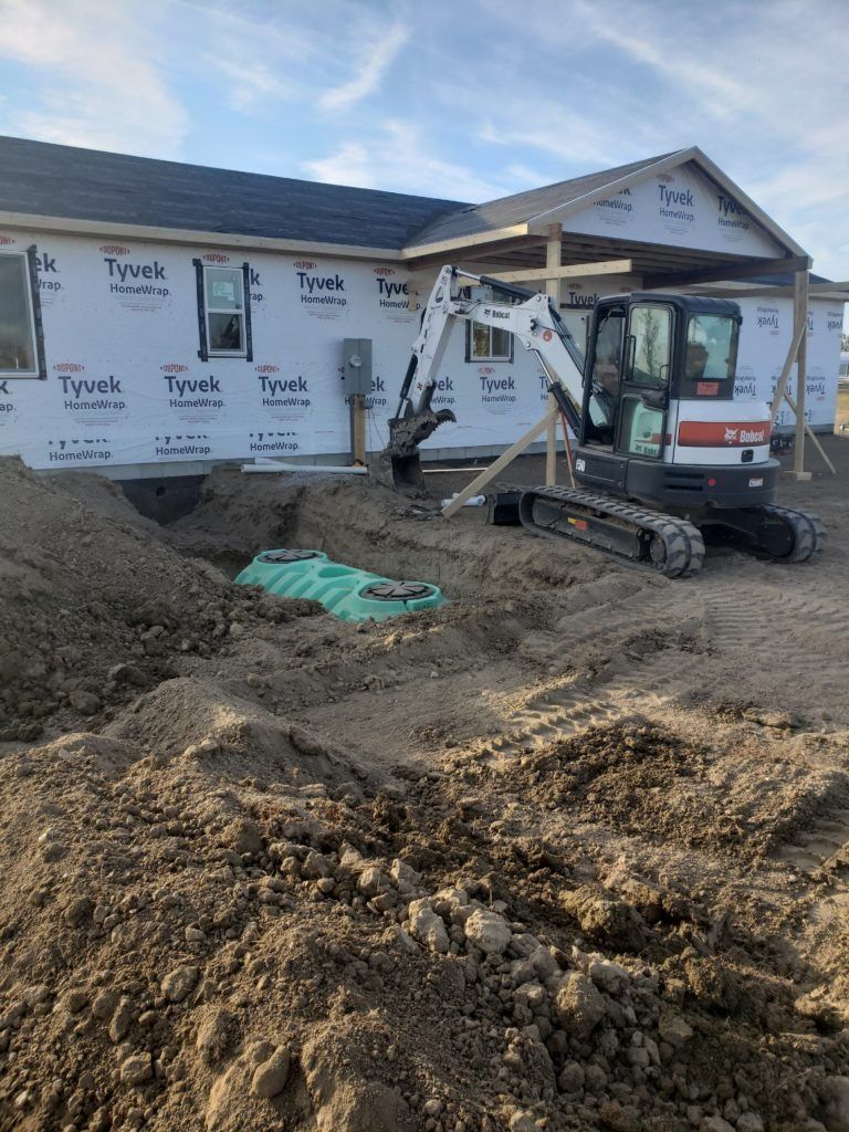 Mini-excavator digging near a house under construction; piles of dirt and a septic tank visible.
