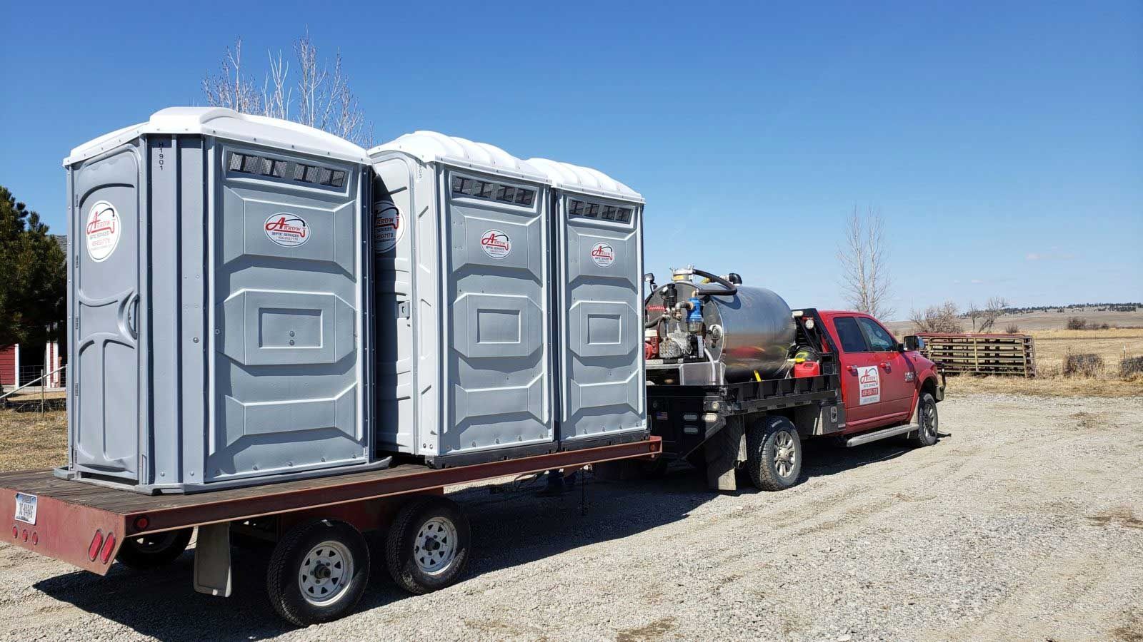 Three portable toilets on a flatbed trailer being pulled by a red truck on a gravel road.
