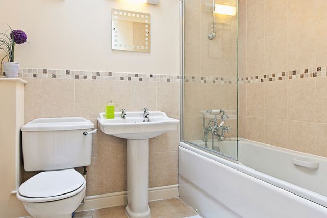 A bathroom featuring a toilet, pedestal sink, and a bathtub with a glass shower screen, set against beige tiled walls.