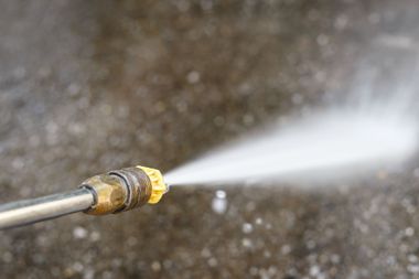 A pressure washer nozzle spraying a high-pressure stream of water onto a dark, textured surface.