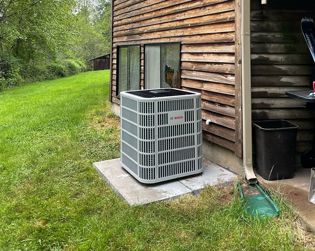 A large air conditioner is sitting in the grass next to a wooden building installed by Ready Energy Solutions.