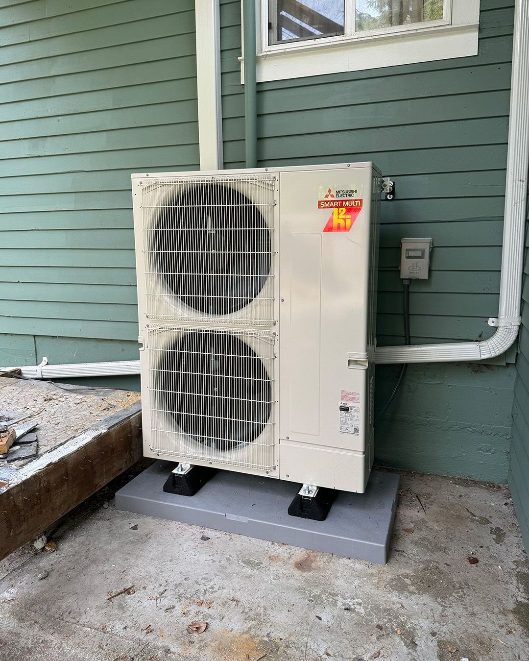 A white air conditioner is sitting on the side of a green house installed by Ready Energy Solutions.