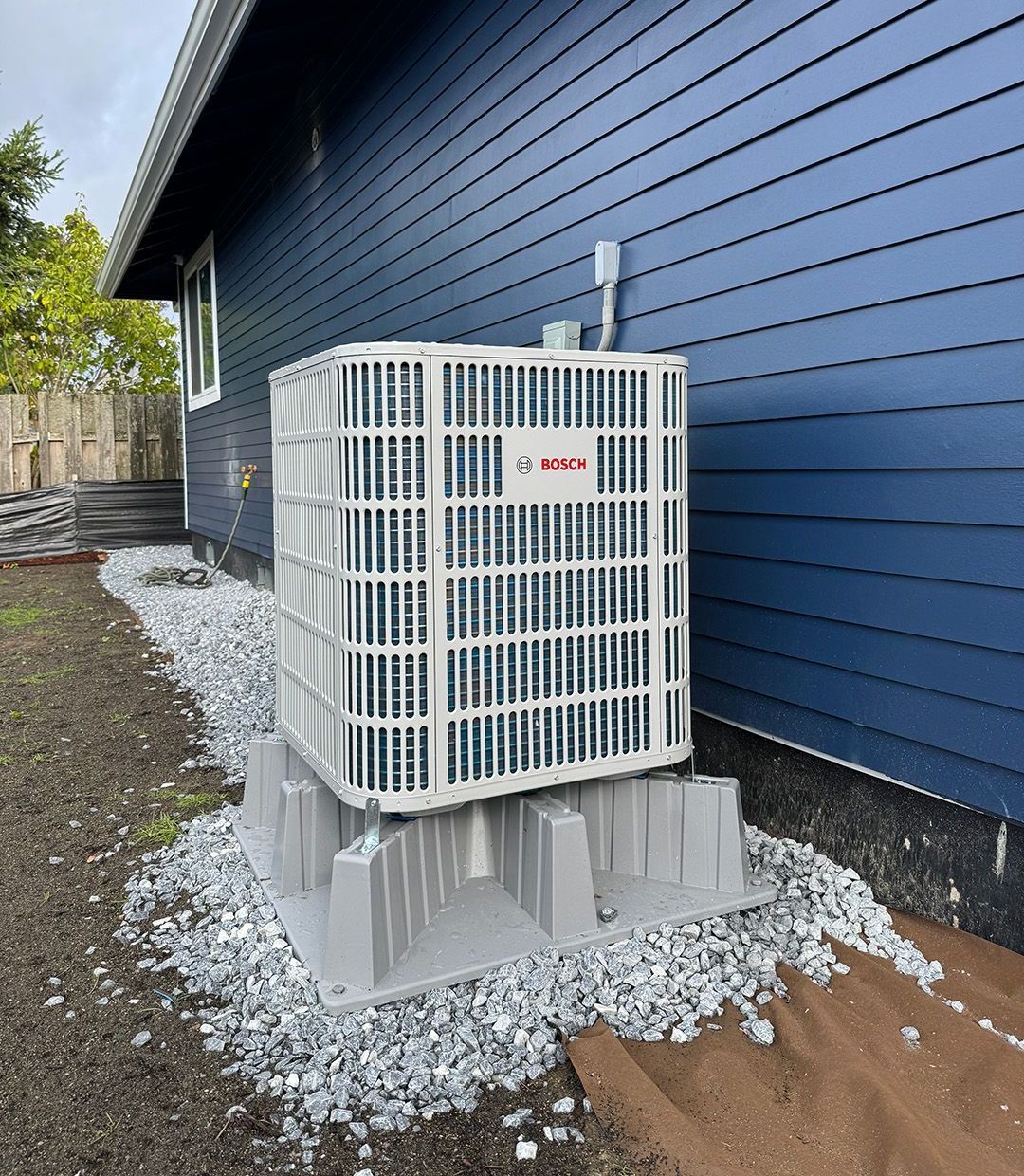 A white air conditioner is sitting on the side of a blue house installed by Ready Energy Solutions.