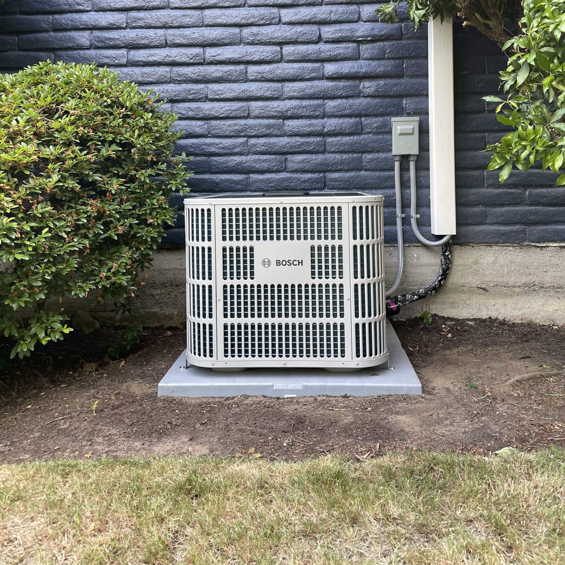 An air conditioner is sitting outside of a house next to a bush.