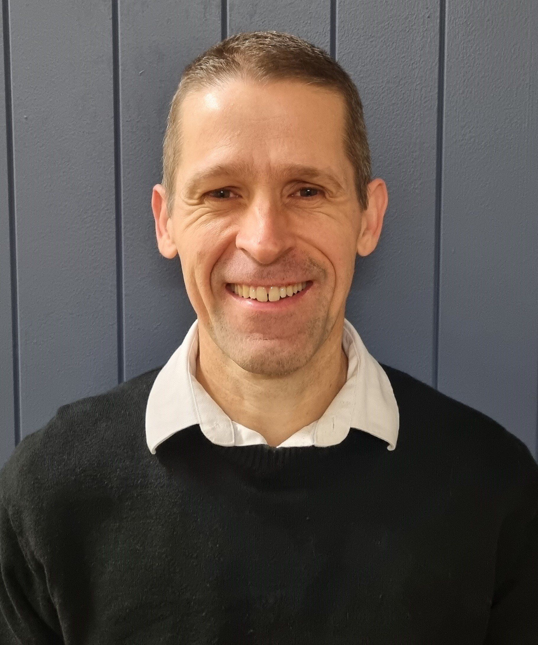 Man with short brown hair and a slight smile, wearing a black sweater and white collared shirt, against a blue wall.