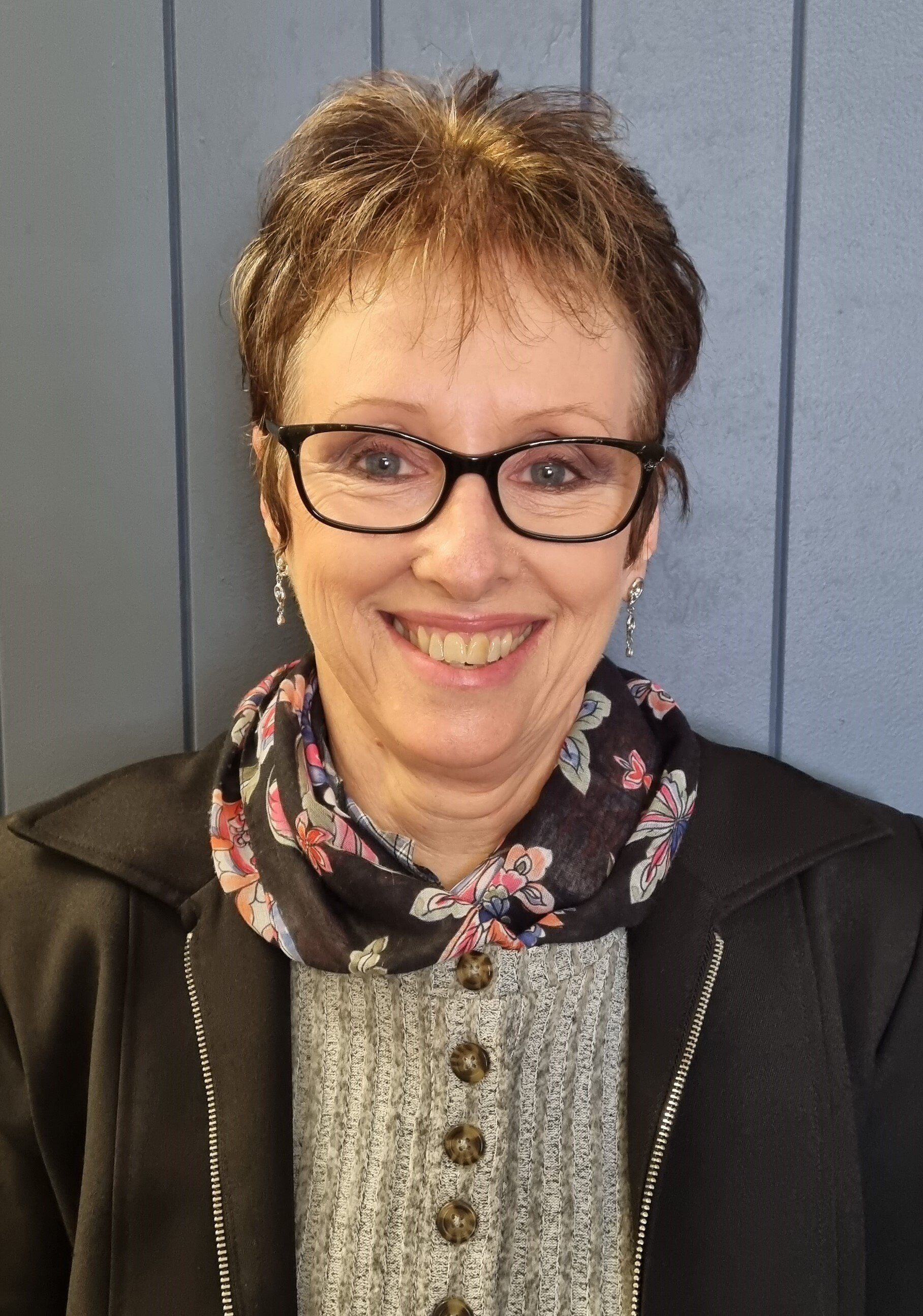 Woman with glasses smiling, wearing a scarf and blazer, in front of a blue wall.