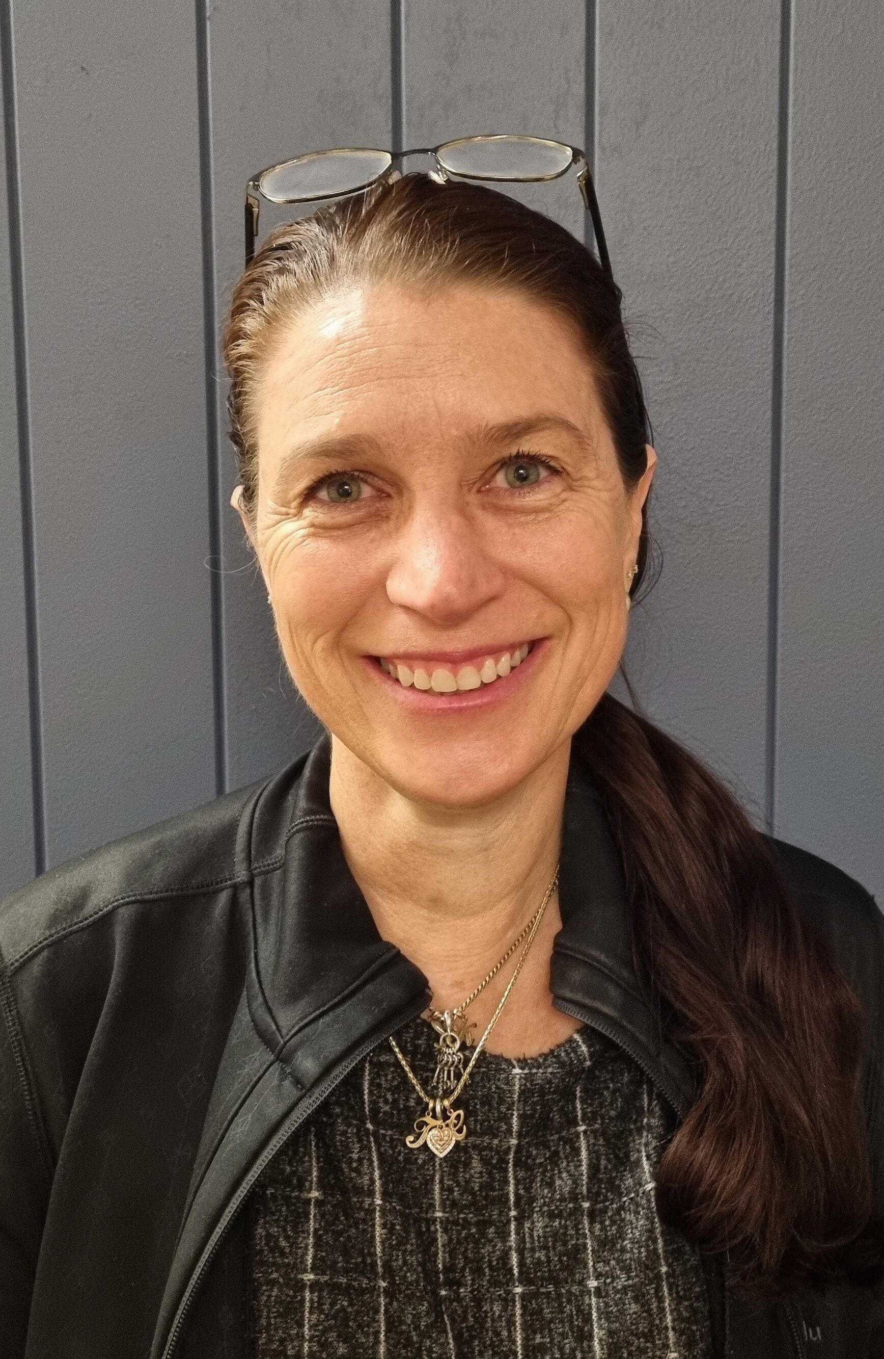 Woman with brown hair smiles, wearing glasses on head, black jacket, and necklace.