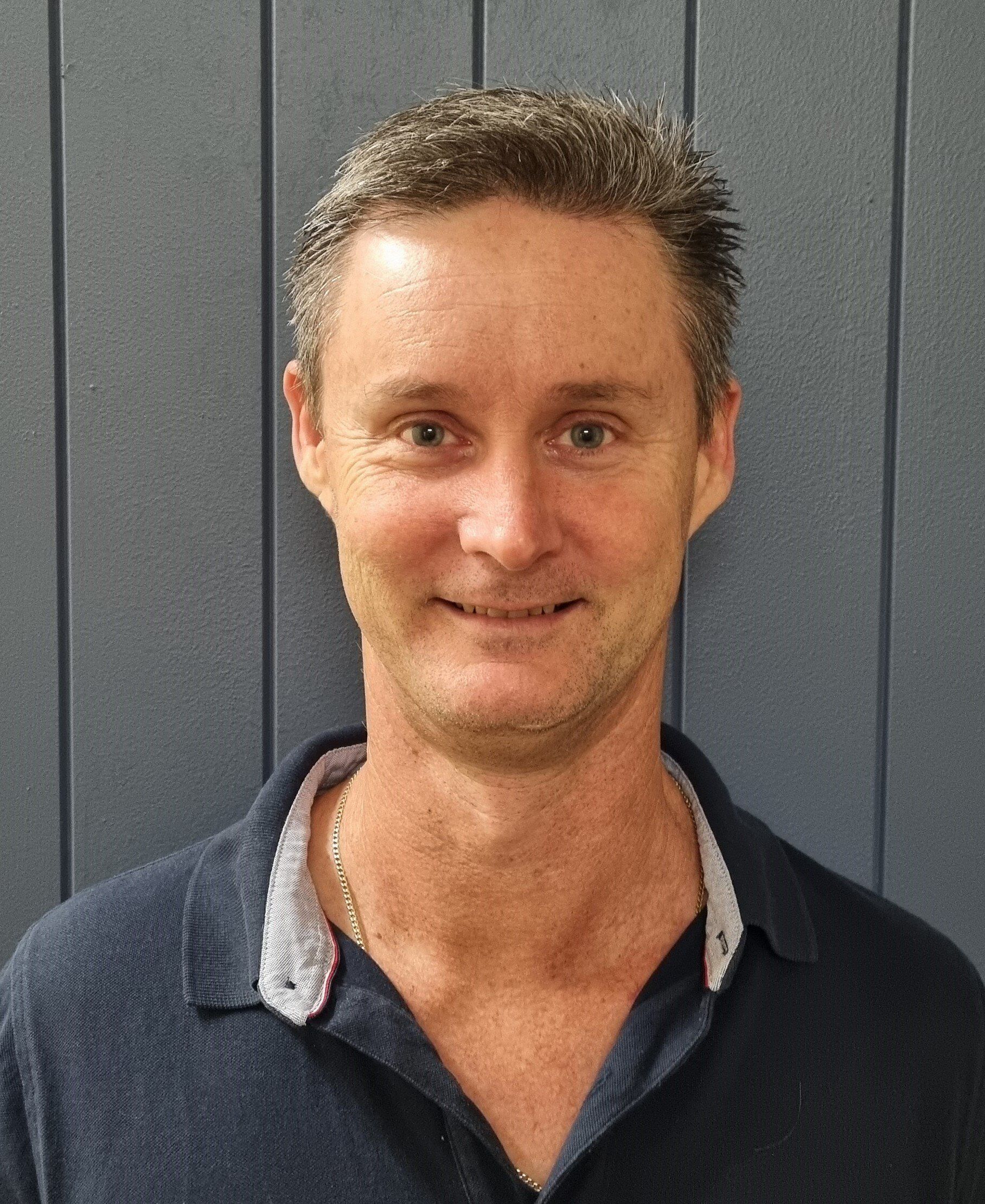 Man with short, graying hair smiling, wearing a dark blue polo shirt. Against a blue wall.