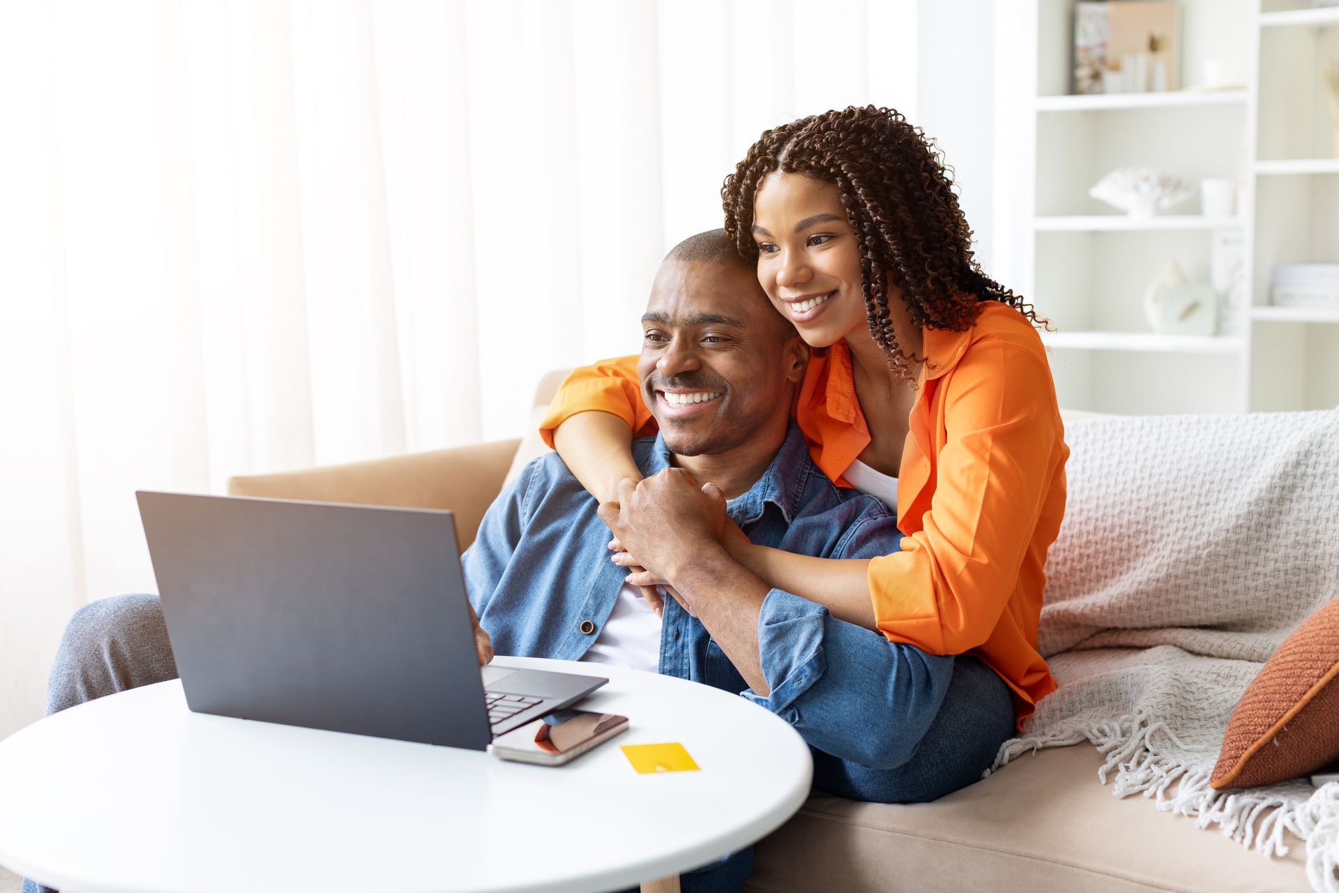 Couple on a couch looking at a laptop. Woman with arm around man, smiling. Bright room with a table.