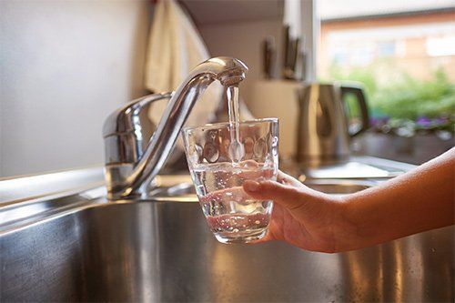 Hand filling a glass with water from a kitchen faucet over a stainless steel sink