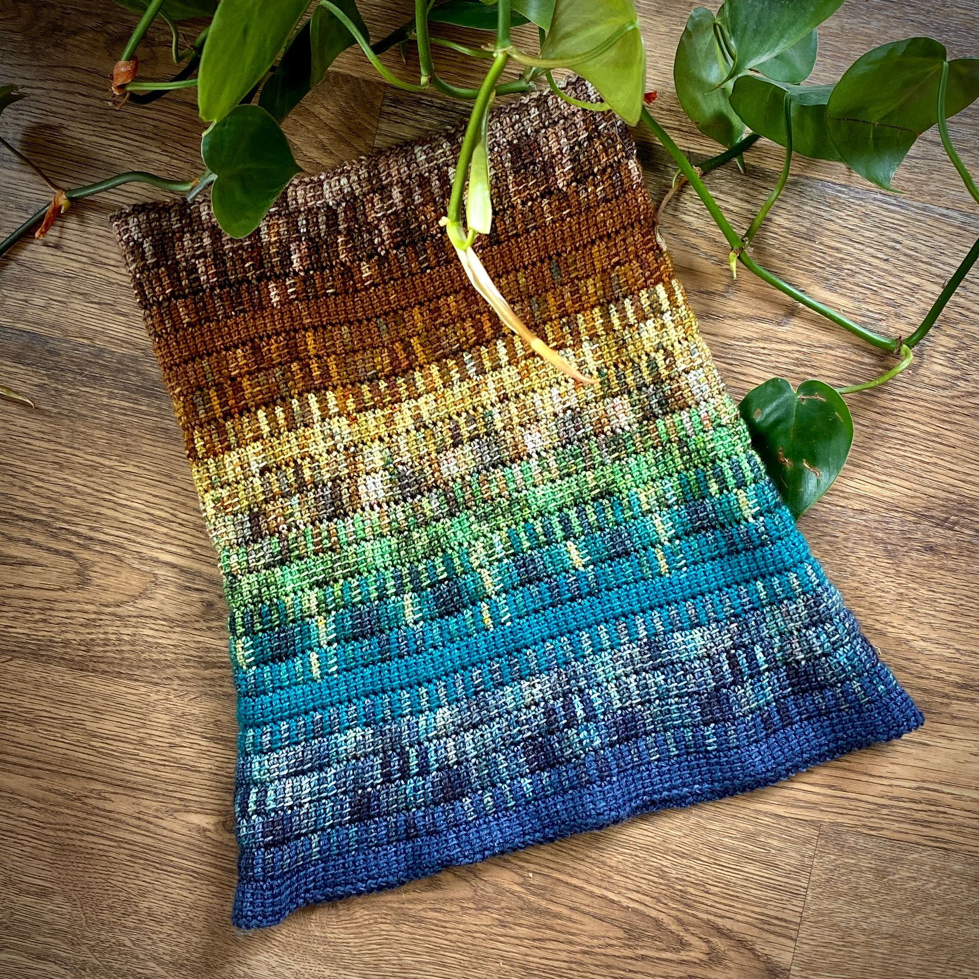Knitted textile with gradient colors: brown to blue, with green plant sprigs on a wooden surface.