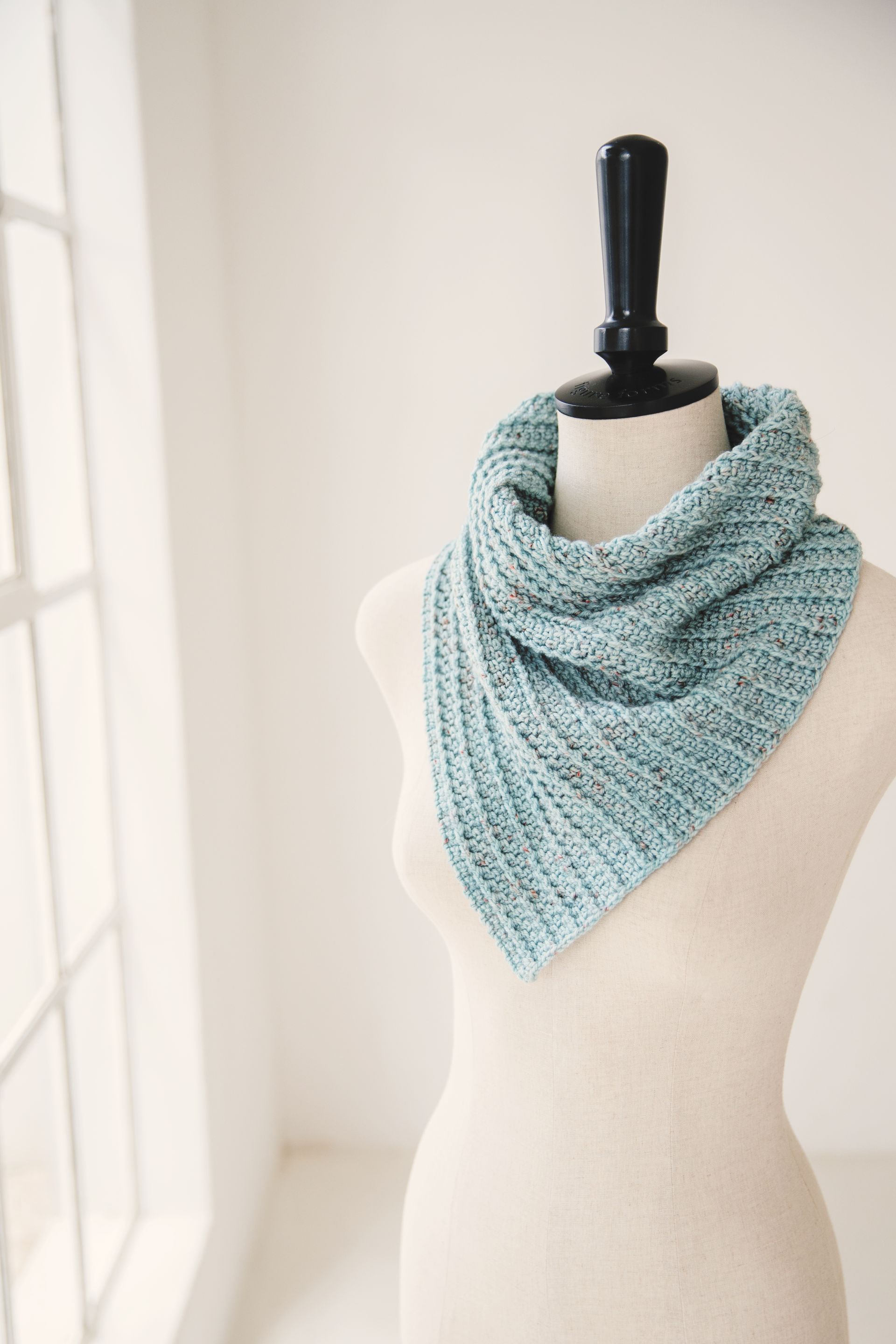 A teal crocheted cowl on a mannequin in front of a window.
