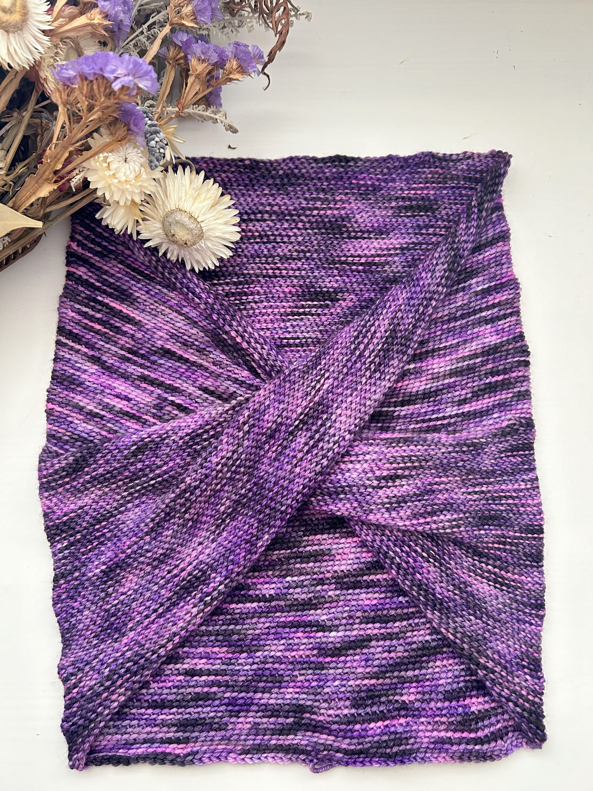Purple and black knit headband with a twisted front, lying on a white surface.