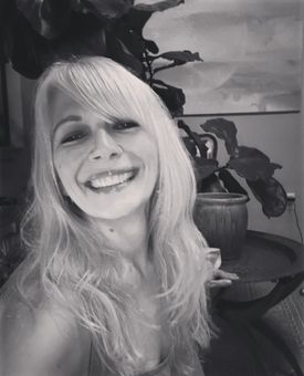 Woman with long blonde hair smiles, indoors near a leafy plant and artwork.