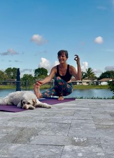 Woman crouching beside a white dog by a lakeside, with blue sky and palm trees in the background
