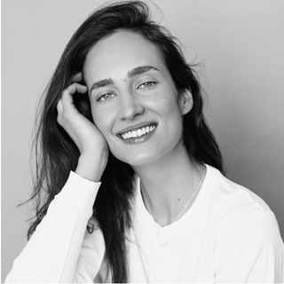 Smiling woman in a white top, resting her head on her hand against a plain background
