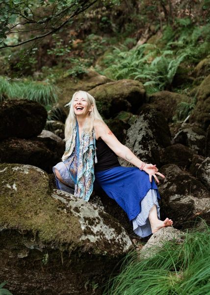 Blonde woman seated on mossy rocks, smiling with arms outstretched. Forest background with ferns.