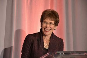 A woman is smiling while sitting at a podium in front of a pink curtain.