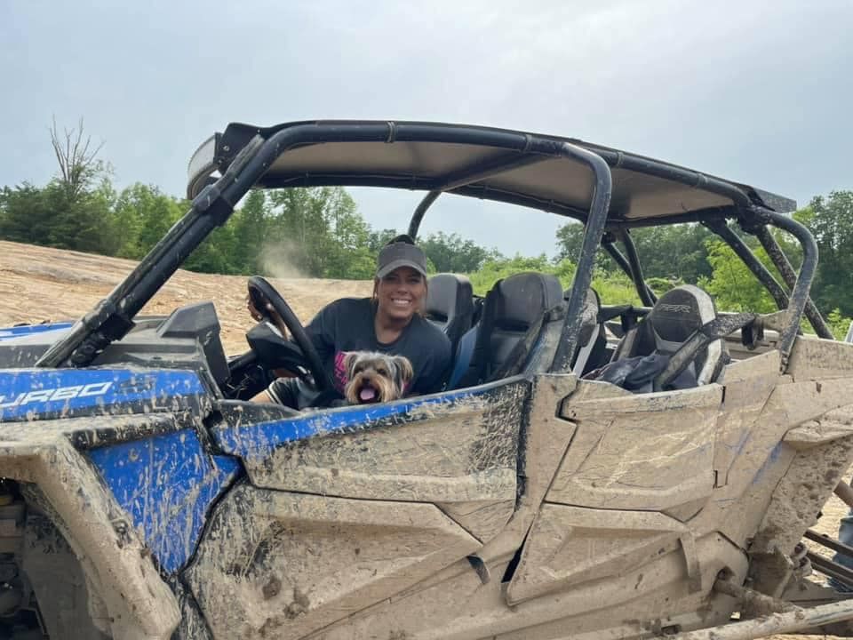 Woman and dog in a muddy ATV, smiling. Off-road setting, blue and white vehicle, sunny day.