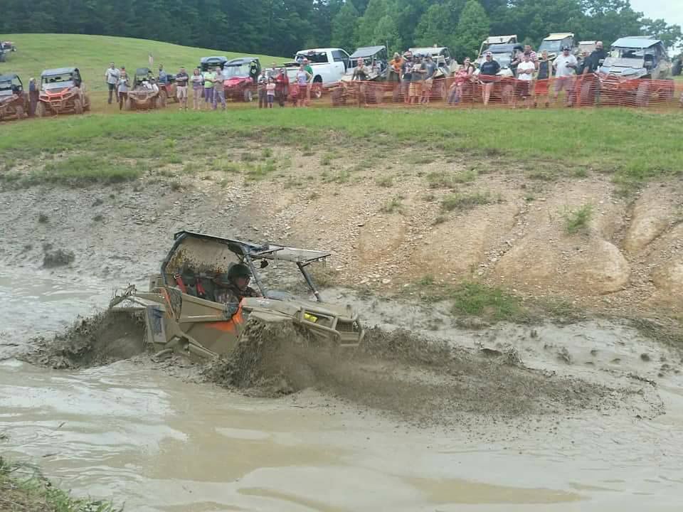 An off-road vehicle struggles in deep mud pit with spectators watching.