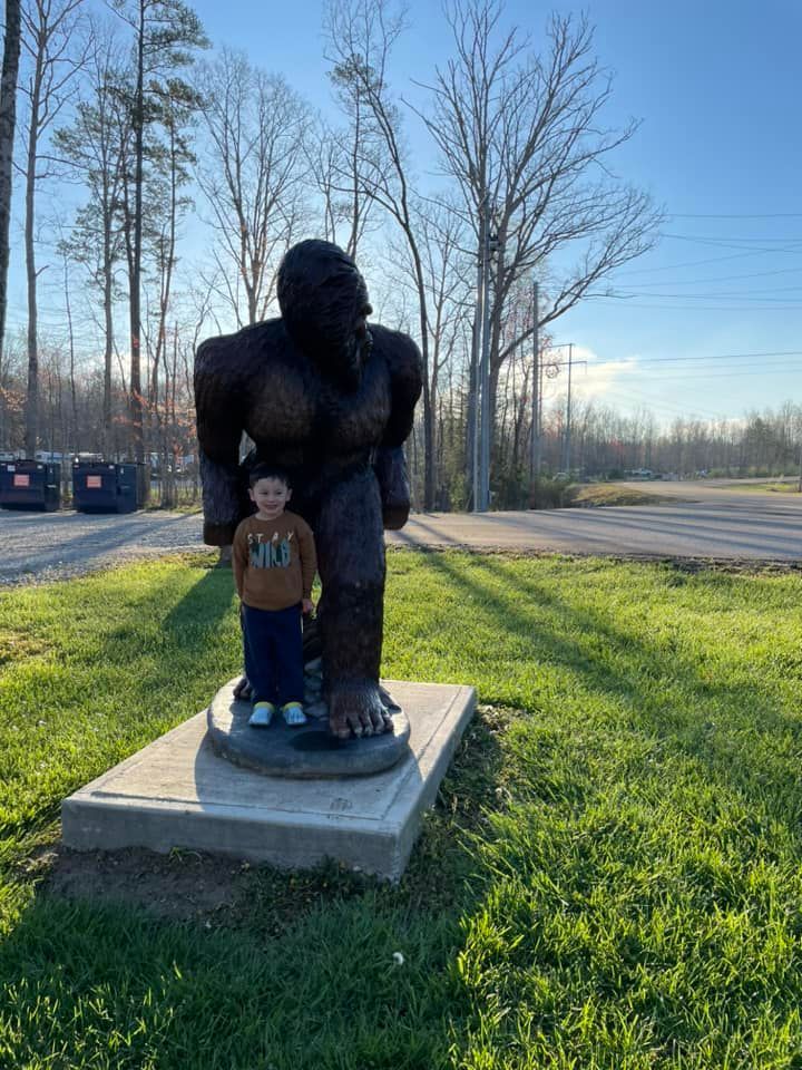 Young child stands next to a large statue of Bigfoot; outdoors, sunny day.