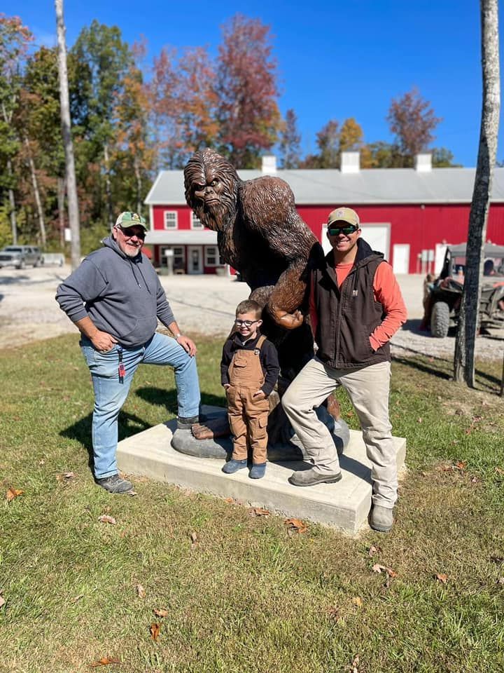 Two men and a child pose with a Bigfoot statue in front of a red building and trees.