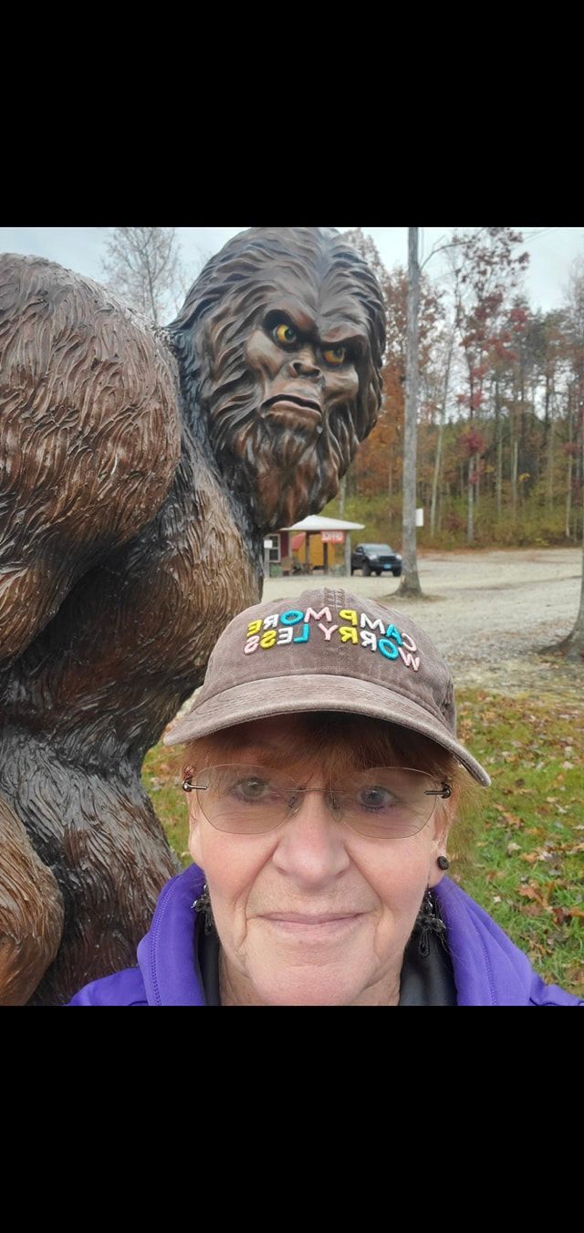 A person smiles in front of a Bigfoot statue. Autumn trees in the background.