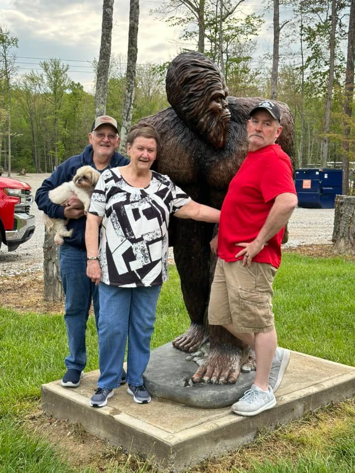 Three people pose with a Bigfoot statue outdoors. One holds a small dog; another rests hand on the statue.