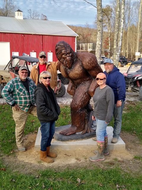 Group of people posing with a Bigfoot statue in front of a red barn and trees.