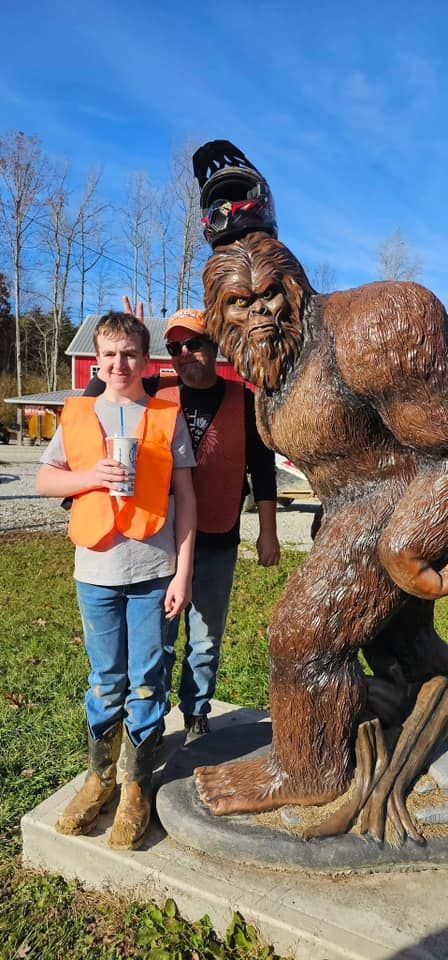 Two people pose with a Bigfoot statue, each wearing orange safety vests, outside on a sunny day.