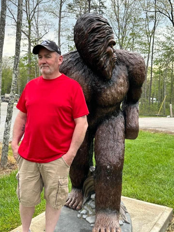 Man in red shirt stands next to a Bigfoot statue outdoors; trees in background.