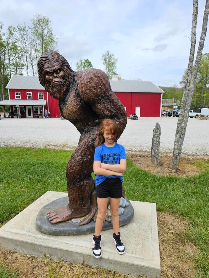 A child poses with arms crossed in front of a Bigfoot statue in a grassy area, with red buildings in the background.