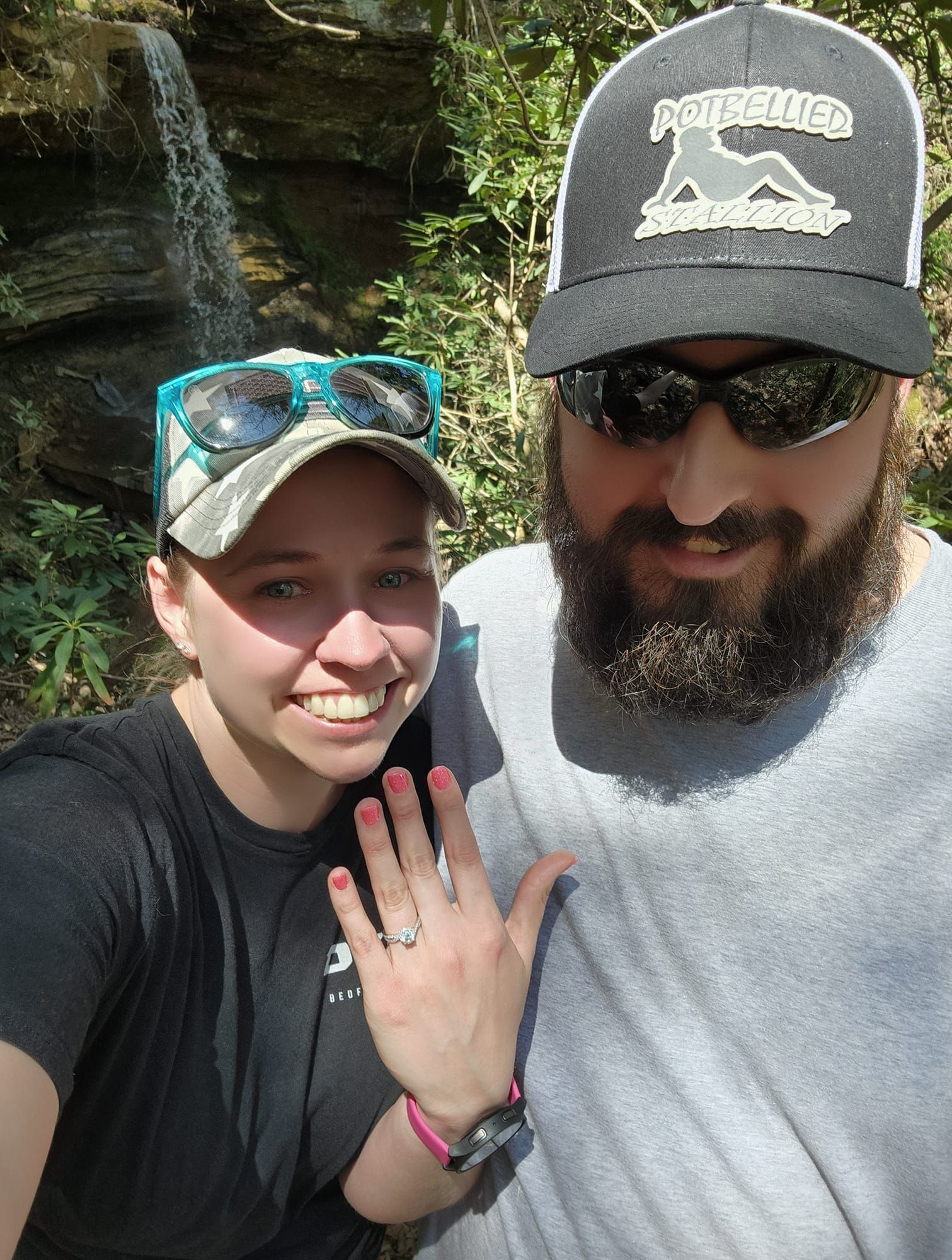 Woman showing engagement ring, smiling. Man in hat, sunglasses, smiling. By waterfall.