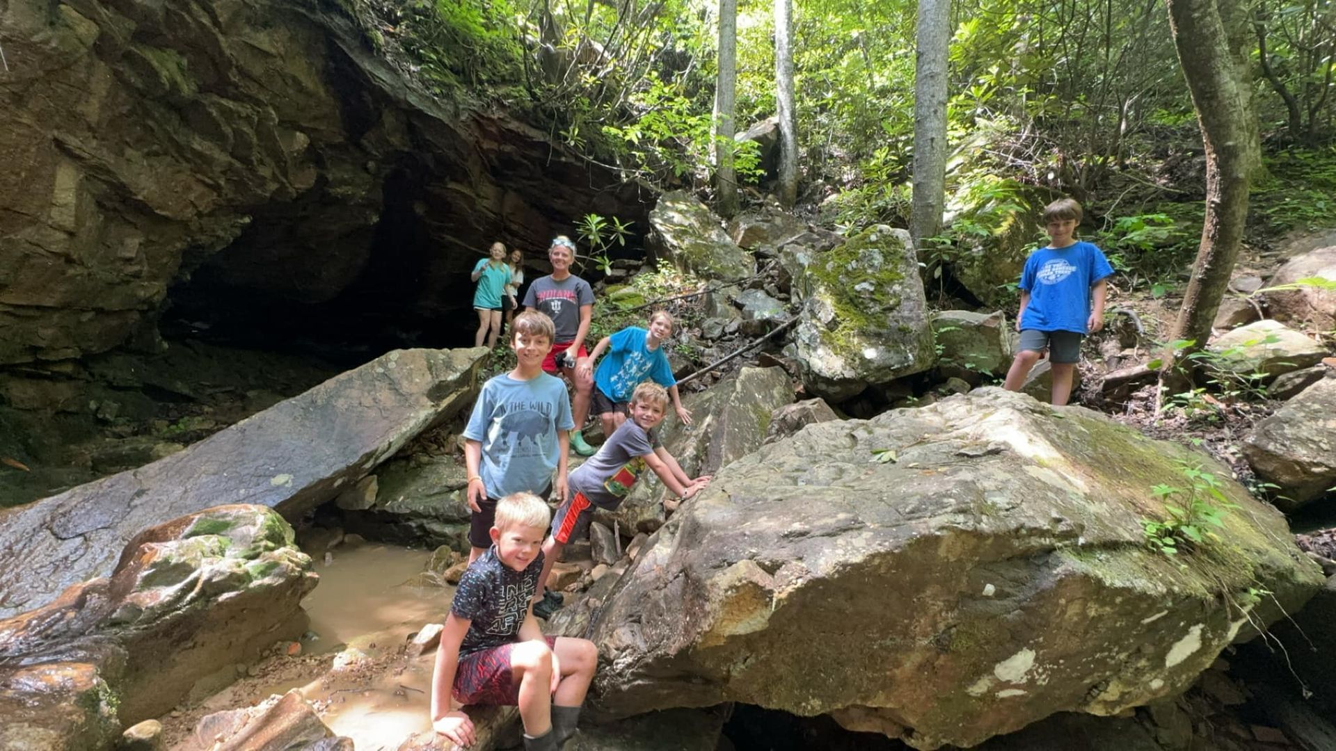 Group of children exploring a rocky, wooded area near a cave.