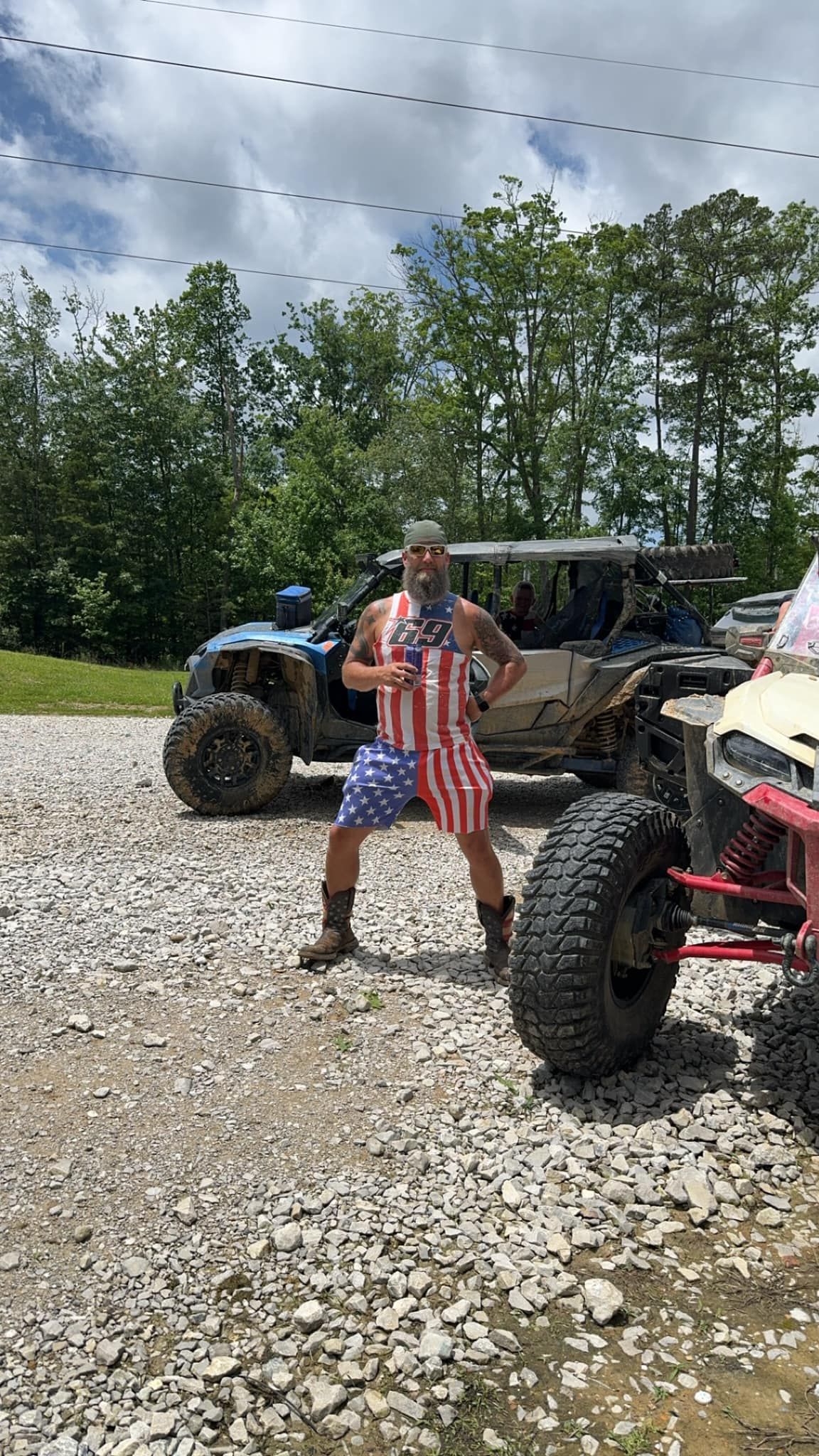 Man in American flag attire poses near off-road vehicles on gravel road.