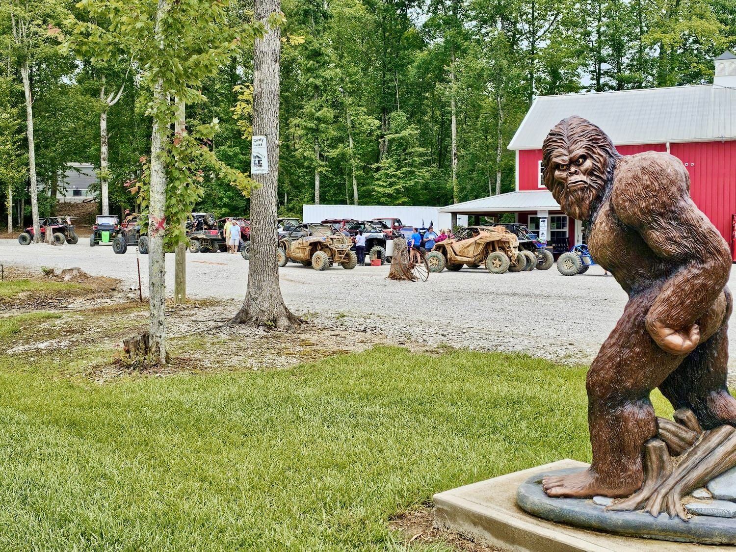 Statue of Bigfoot stands in front of a red building and parked ATVs on a gravel lot; trees in background.