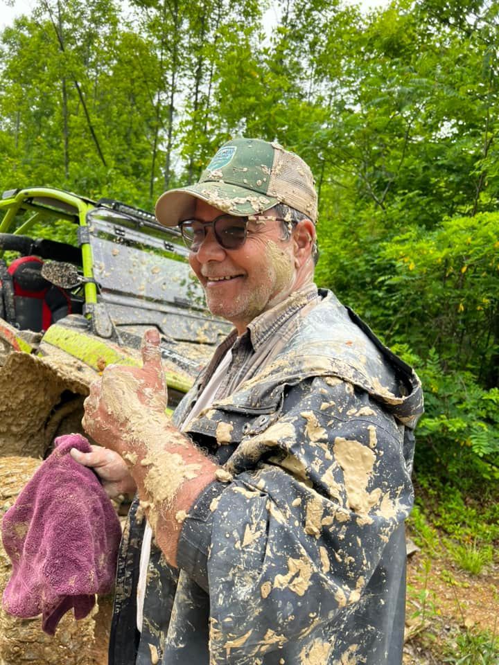 Man covered in mud gives thumbs up next to an ATV, green trees in background.