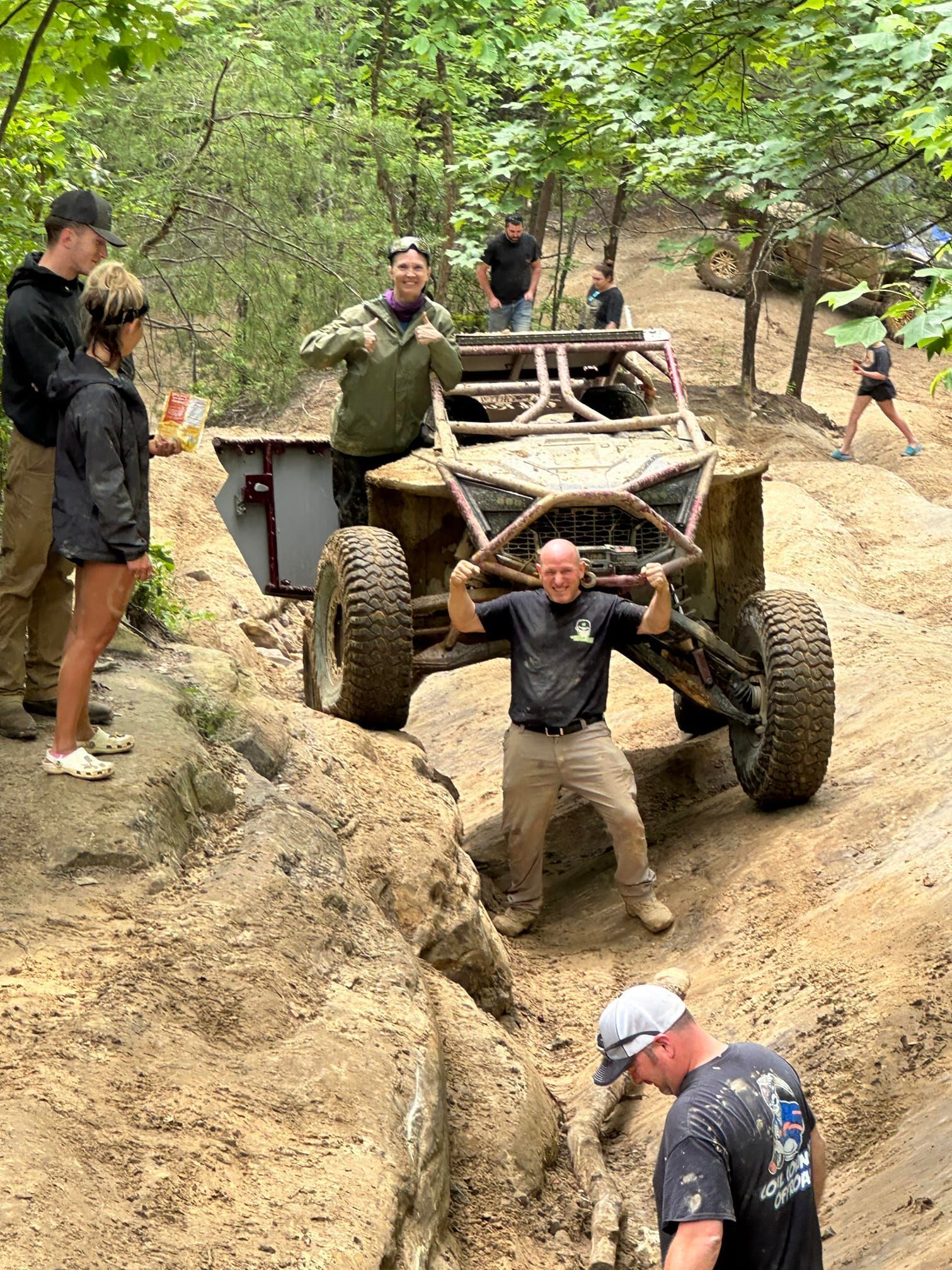 Off-road vehicle ascends a steep dirt incline; people cheer and watch in a wooded area.