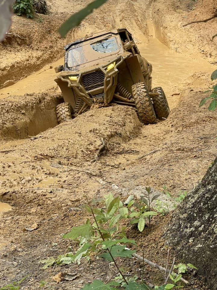 Muddy UTV climbs a steep, muddy hill; tan vehicle, brown mud, green plants.