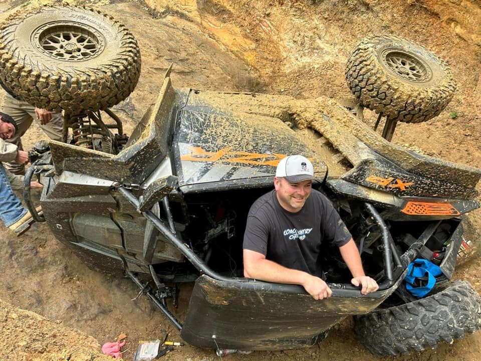 Man smiling from rolled-over off-road vehicle in mud. A person assists, outdoor setting.