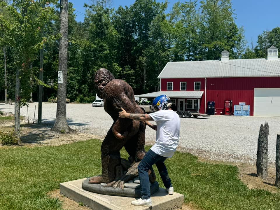 A person pretends to wrestle a Bigfoot statue outside a red building on a sunny day.