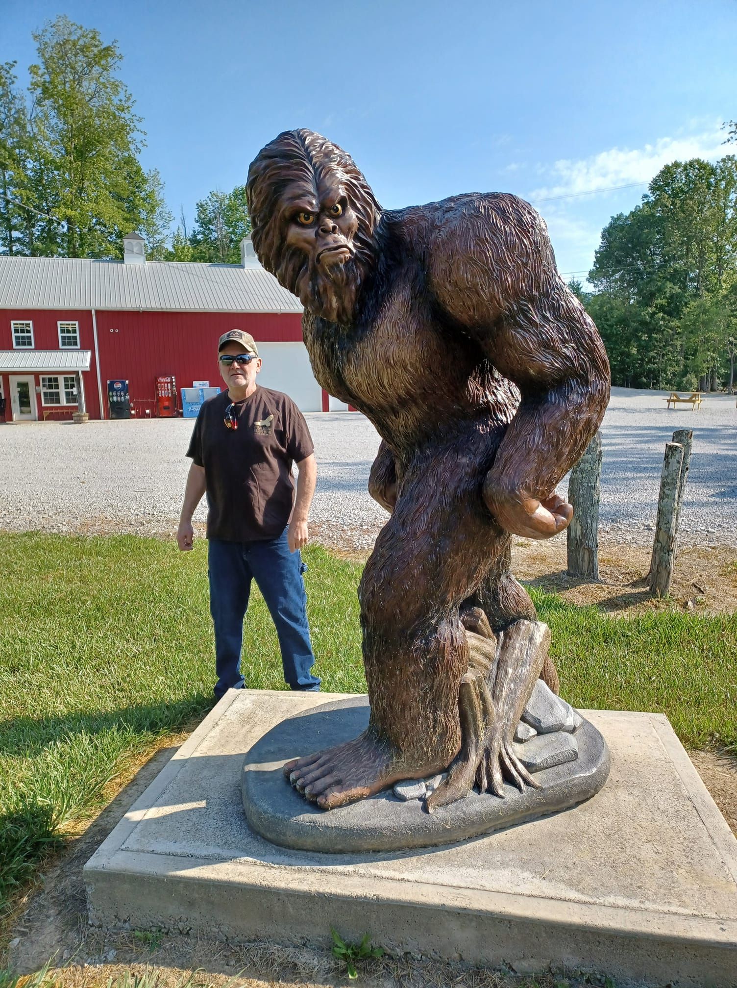 Man stands next to a large Bigfoot statue. The statue is brown and detailed, on a concrete base, in front of a red building.