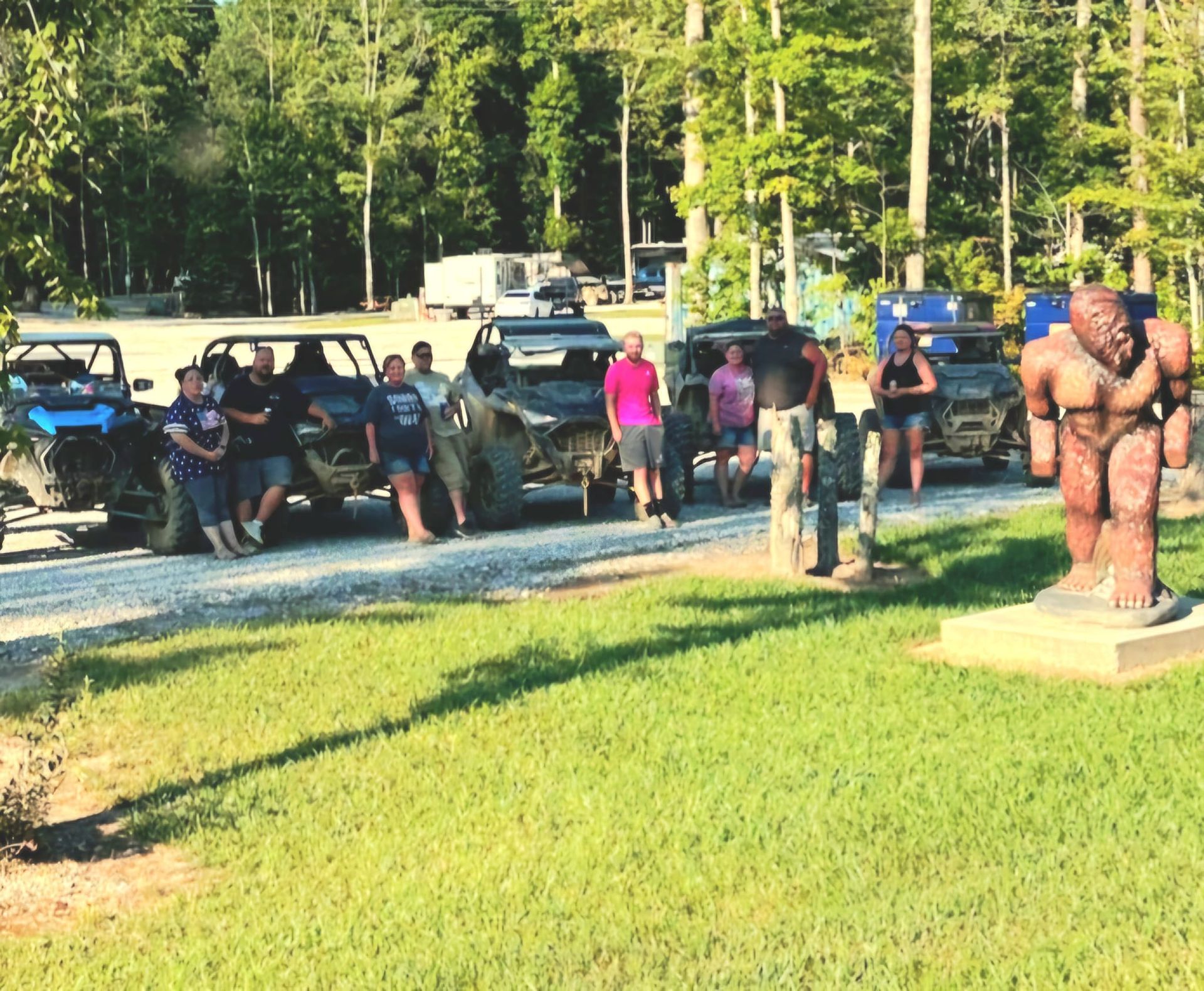 People pose with ATVs near a Bigfoot statue on a sunny day.