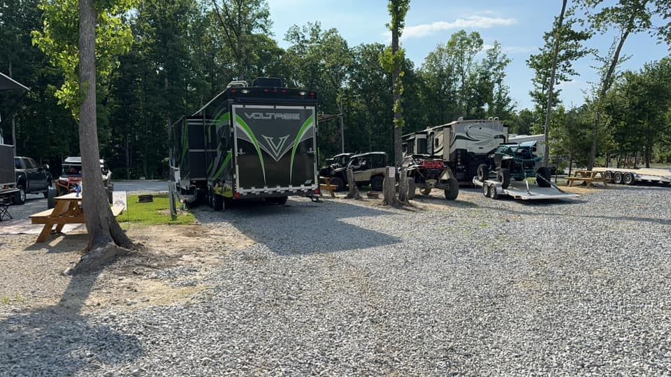RV campsite with trailers and vehicles. Gravel ground, trees, sunny day.