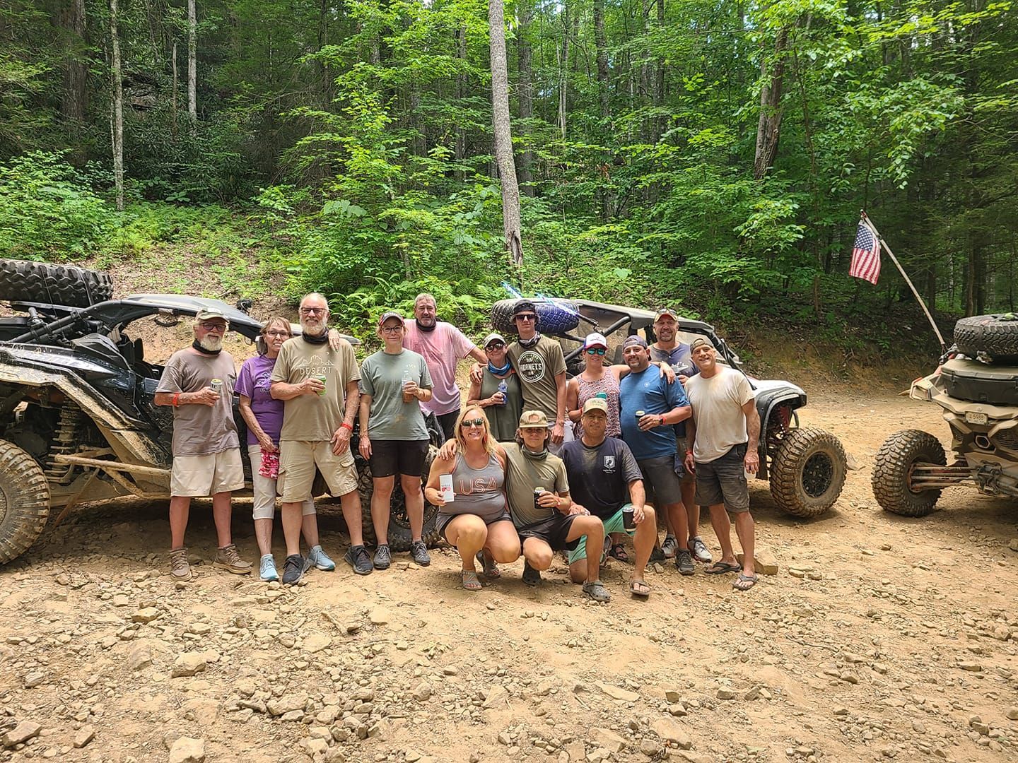 Group of people posing with off-road vehicles in a wooded area. They are smiling.